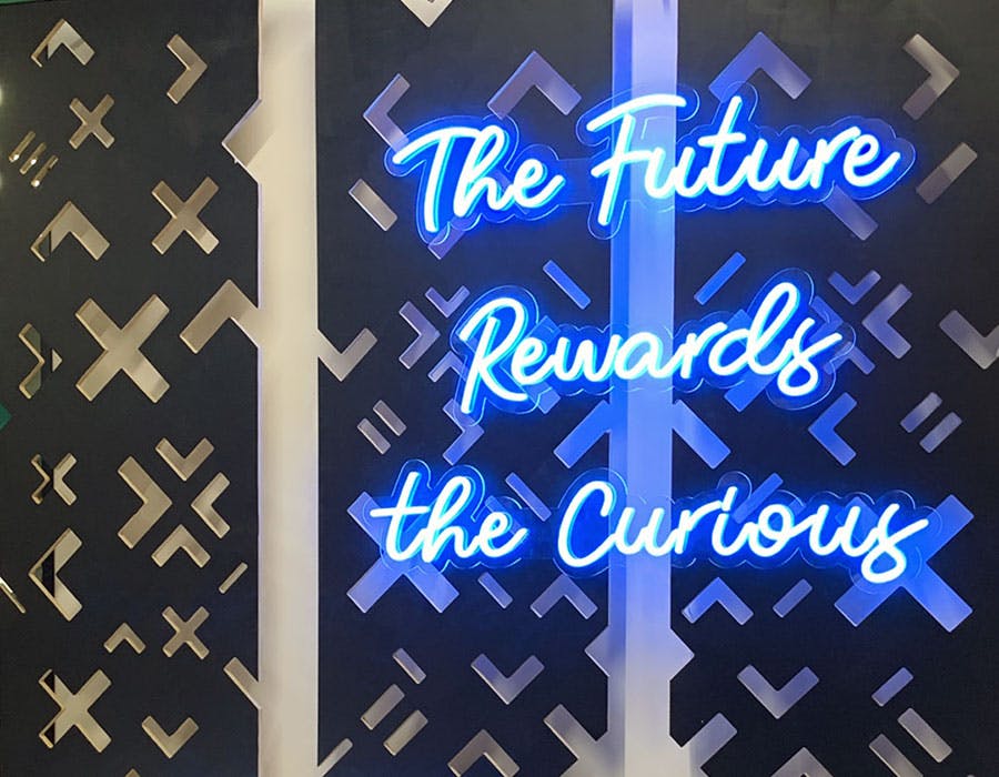 Blue neon sign on black and white wall reads “the future rewards the curious”