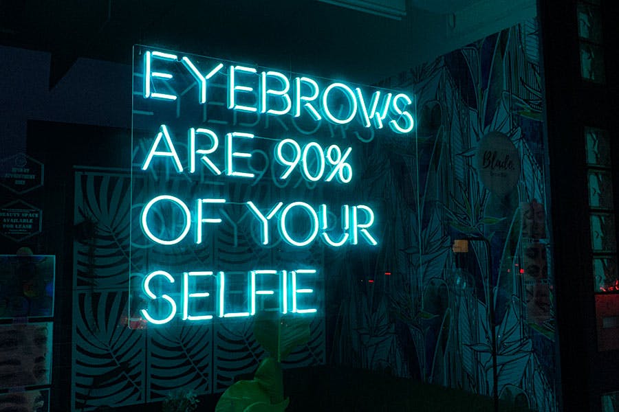 teal neon sign reads eyebrows are 90% of your selfie